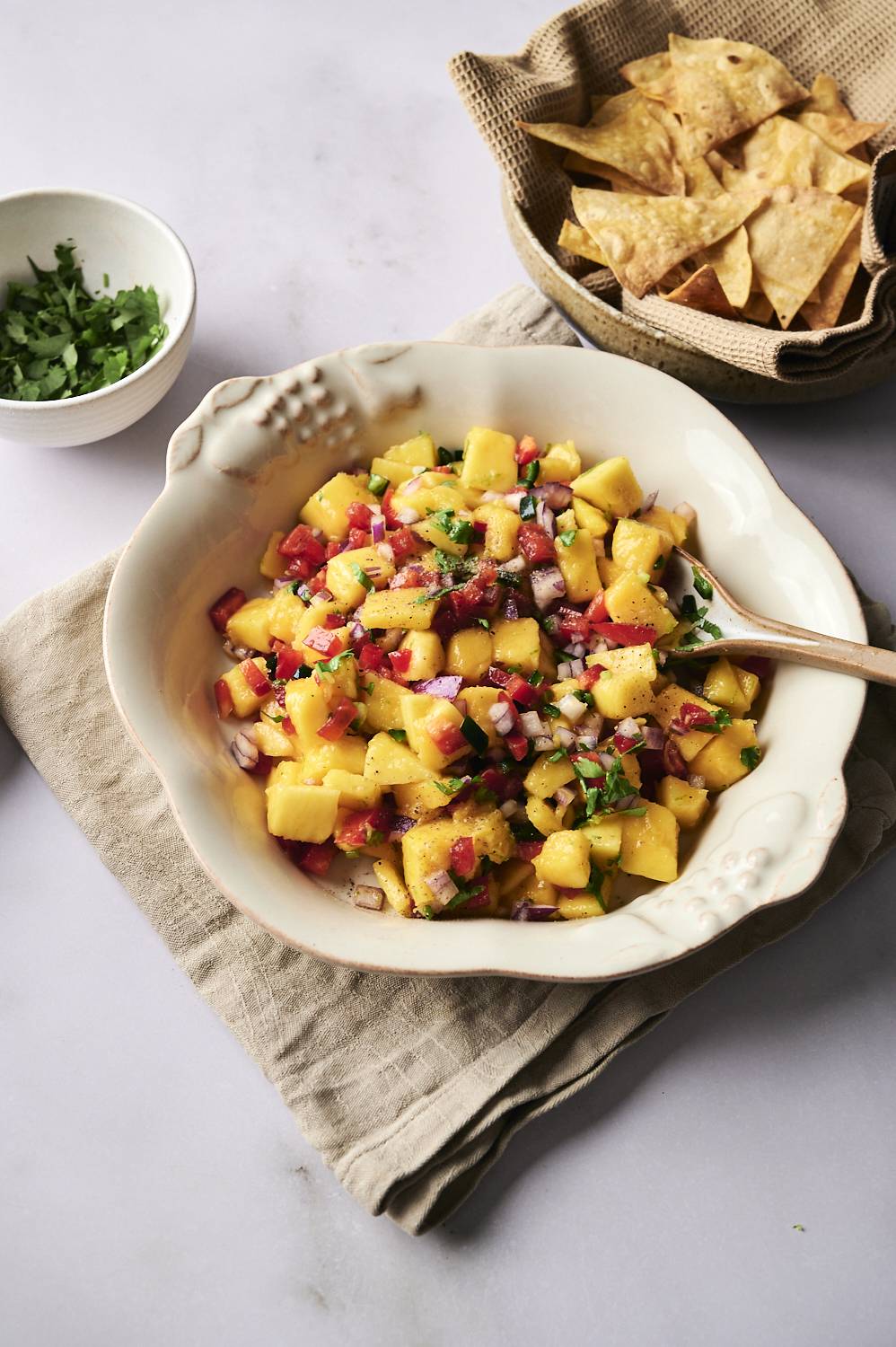 Fresh mango salsa in a white bowl with a wooden spoon, featuring diced mango, red onion, red bell pepper, jalapeño, and cilantro, served with a side of tortilla chips.