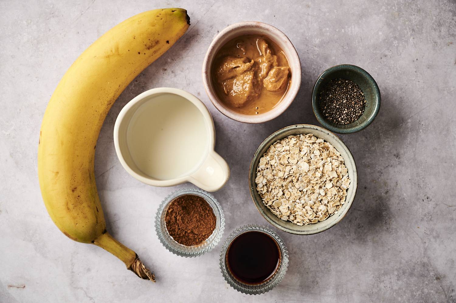 A top-down shot of the ingredients for overnight oatmeal, including jars with oats and chia seeds, a banana, and a jar of peanut butter.