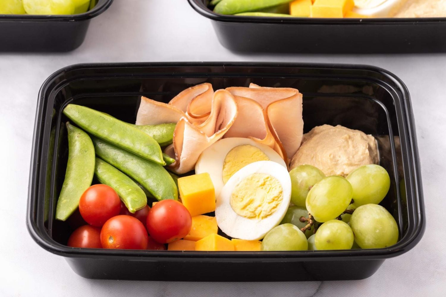 Hard boiled egg protein box with boiled eggs, hummus, grapes, cheese, hummus, and vegetables.