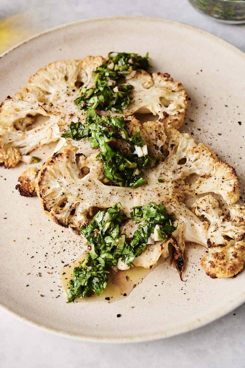 Roasted cauliflower steaks plated with vibrant green chimichurri sauce and cracked black pepper.