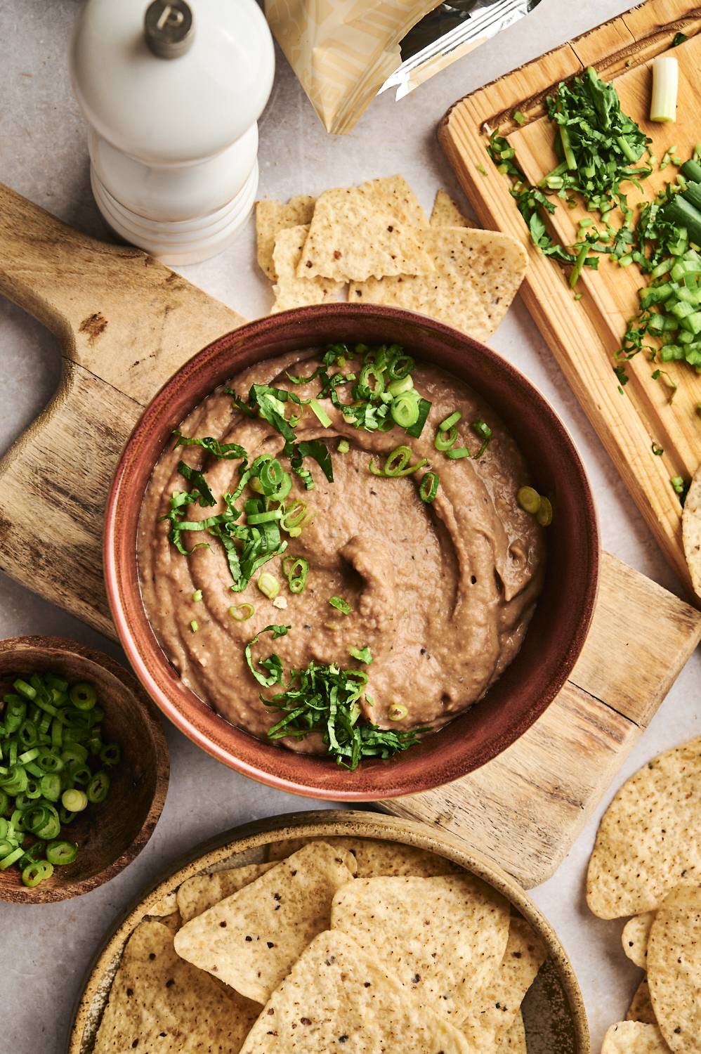 A bowl of creamy refried beans garnished with green onions and cilantro is served on a wooden board alongside tortilla chips and fresh toppings.