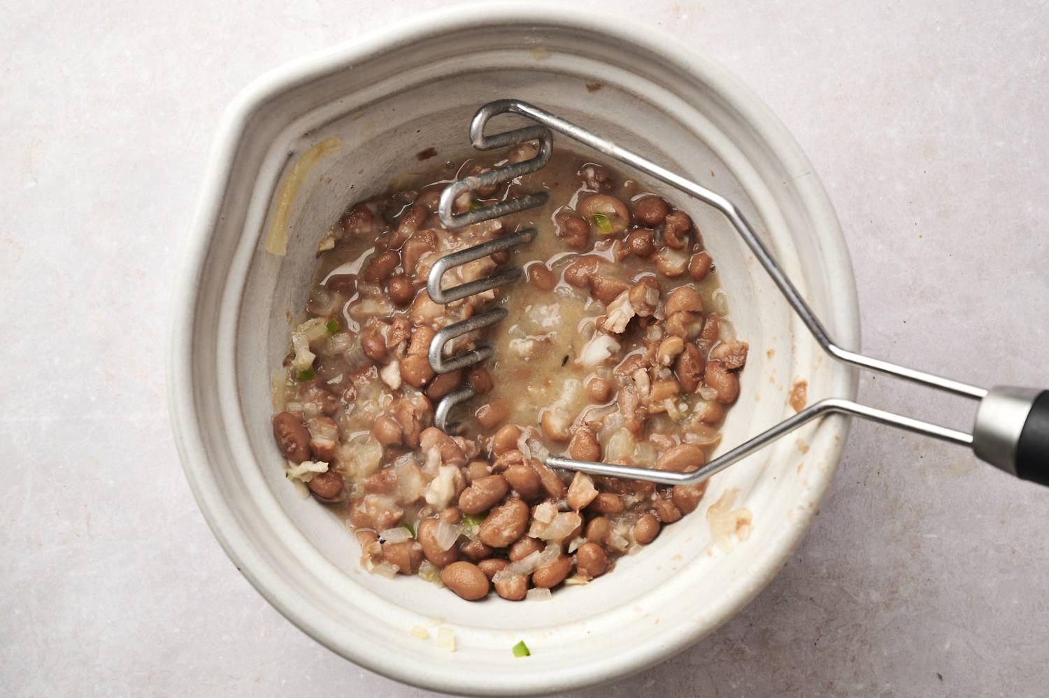 Cooked pinto beans are being mashed in a bowl with a potato masher, creating a smooth and creamy texture.