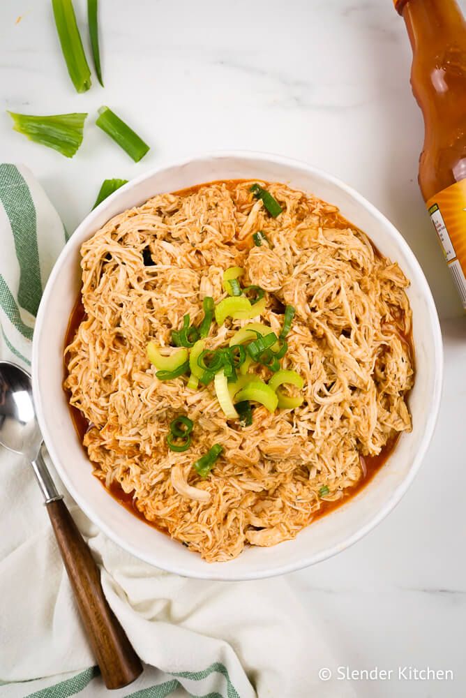 Buffalo chicken that was cooked in a slow cooker and shredded in a bowl with a wooden spoon, celery, and green onions.