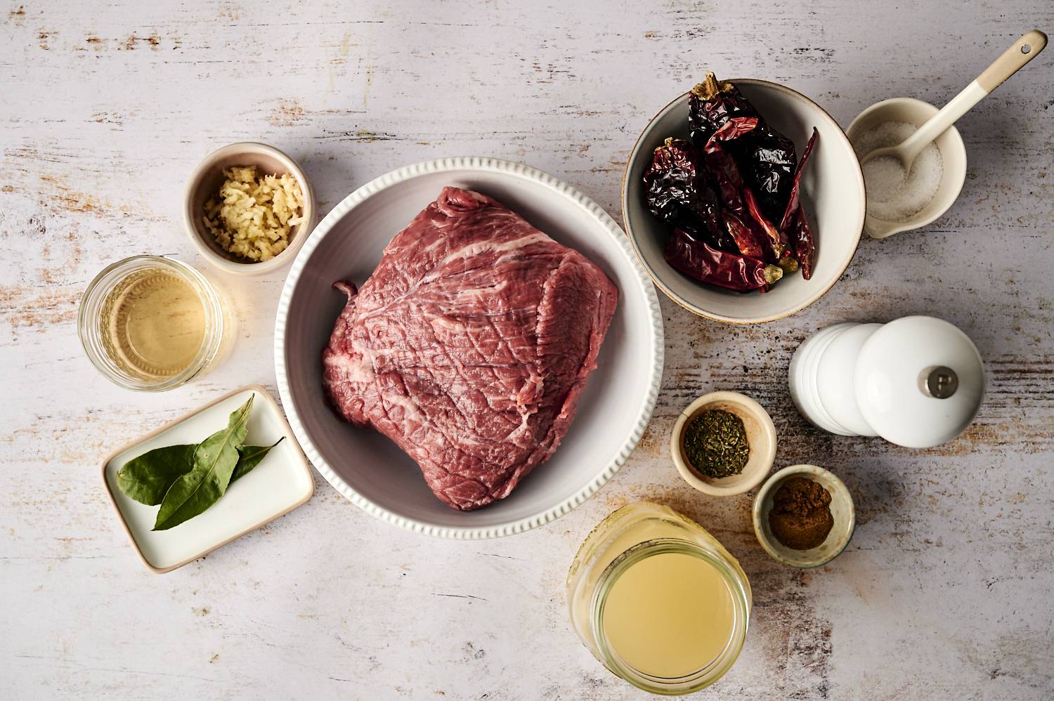 A raw beef roast is surrounded by ingredients for Mexican beef adobo, including dried chilies, garlic, bay leaves, spices, broth, and vinegar, ready for slow cooking.