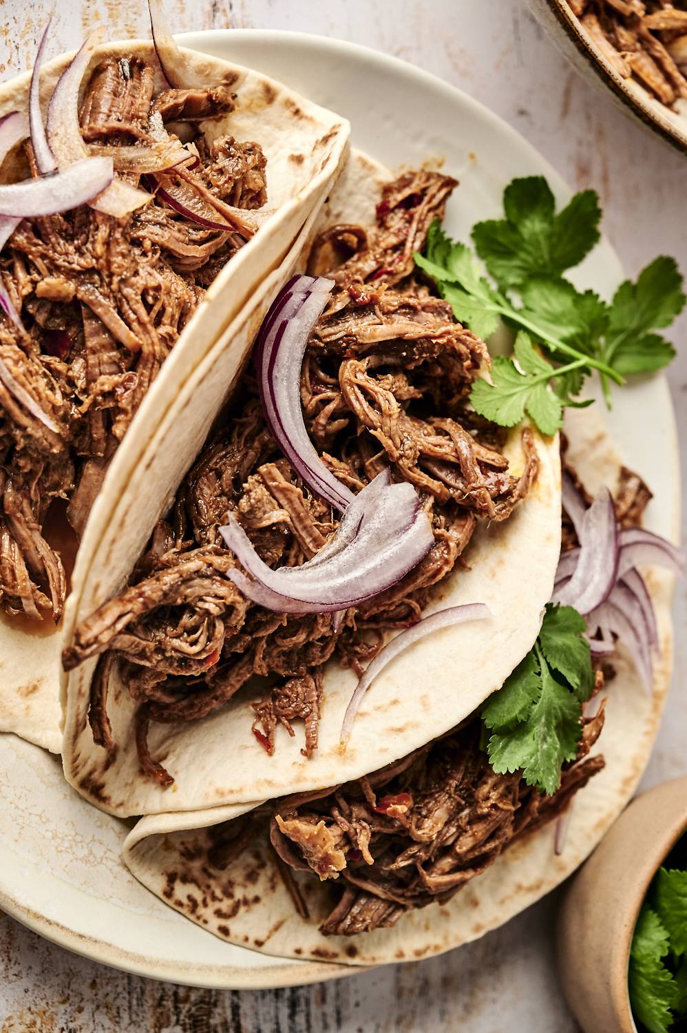 Soft flour tortillas are filled with shredded Mexican beef adobo, topped with sliced red onions and fresh cilantro for a flavorful and hearty meal.