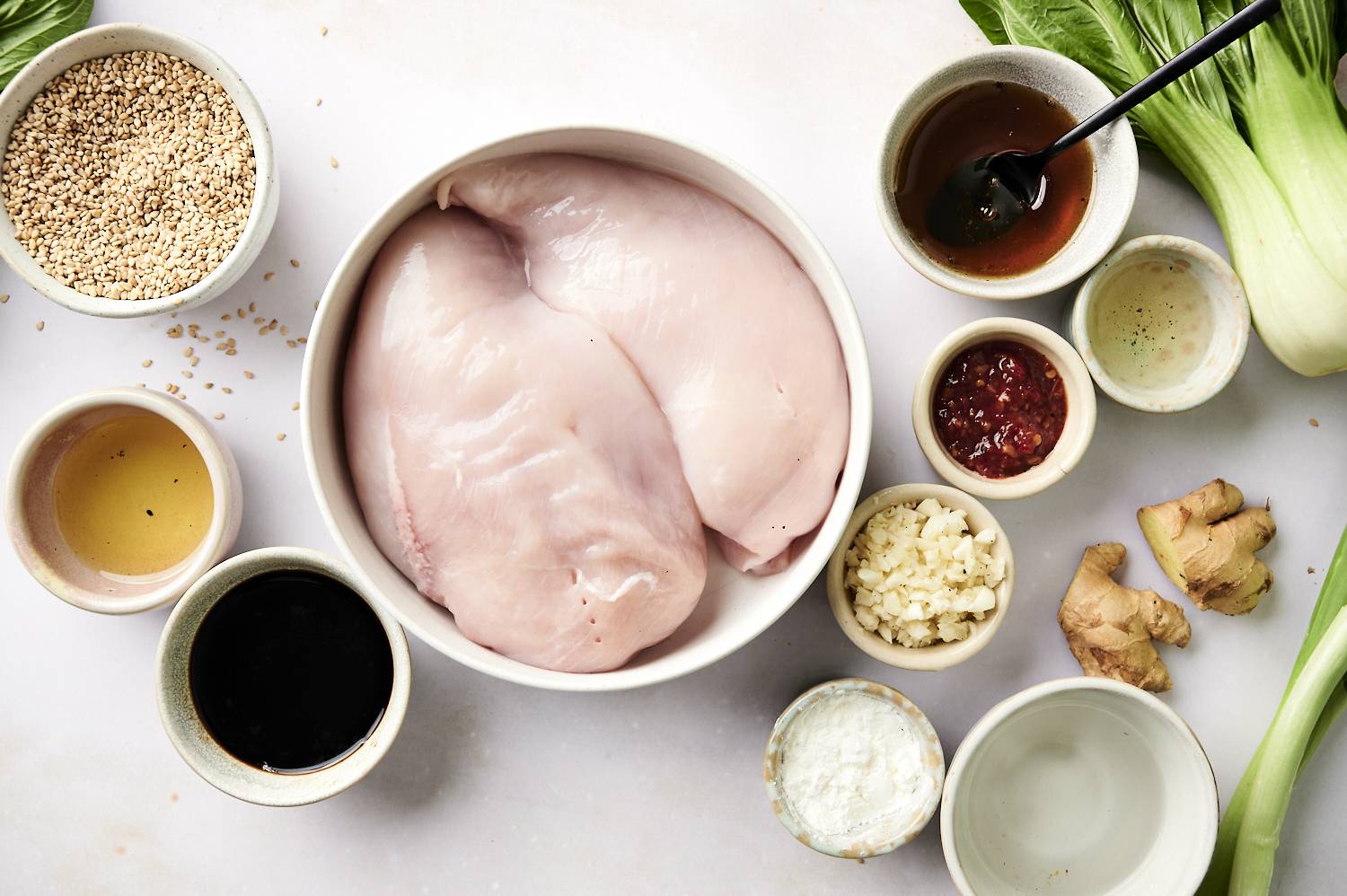 Raw chicken breasts surrounded by individual bowls containing sesame seeds, soy sauce, minced garlic, fresh ginger, chili paste, honey, cornstarch, and sesame oil, with fresh bok choy on the side.