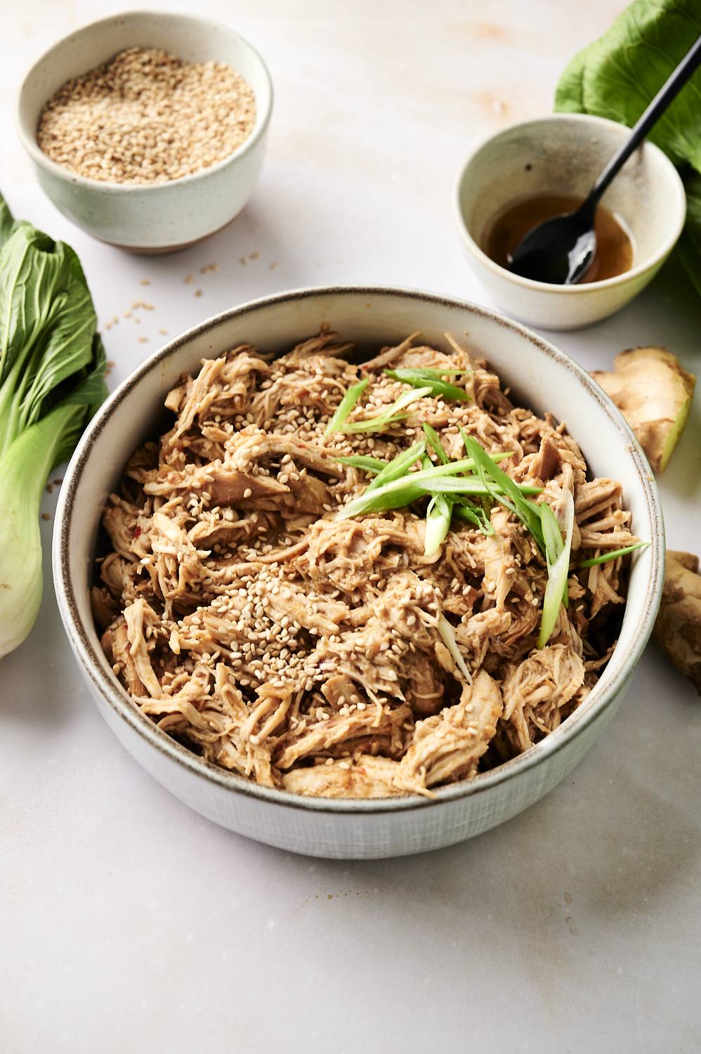 A bowl of shredded slow-cooked sesame ginger chicken garnished with sesame seeds and green onions, surrounded by sesame seeds, bok choy, and a small bowl of sauce in the background.