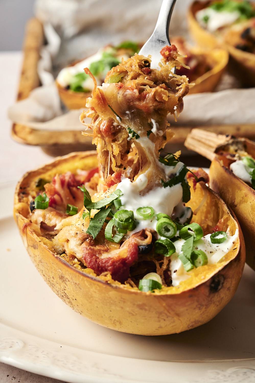 A fork lifting a bite of cheesy spaghetti squash enchilada boat topped with sour cream, green onions, and cilantro.