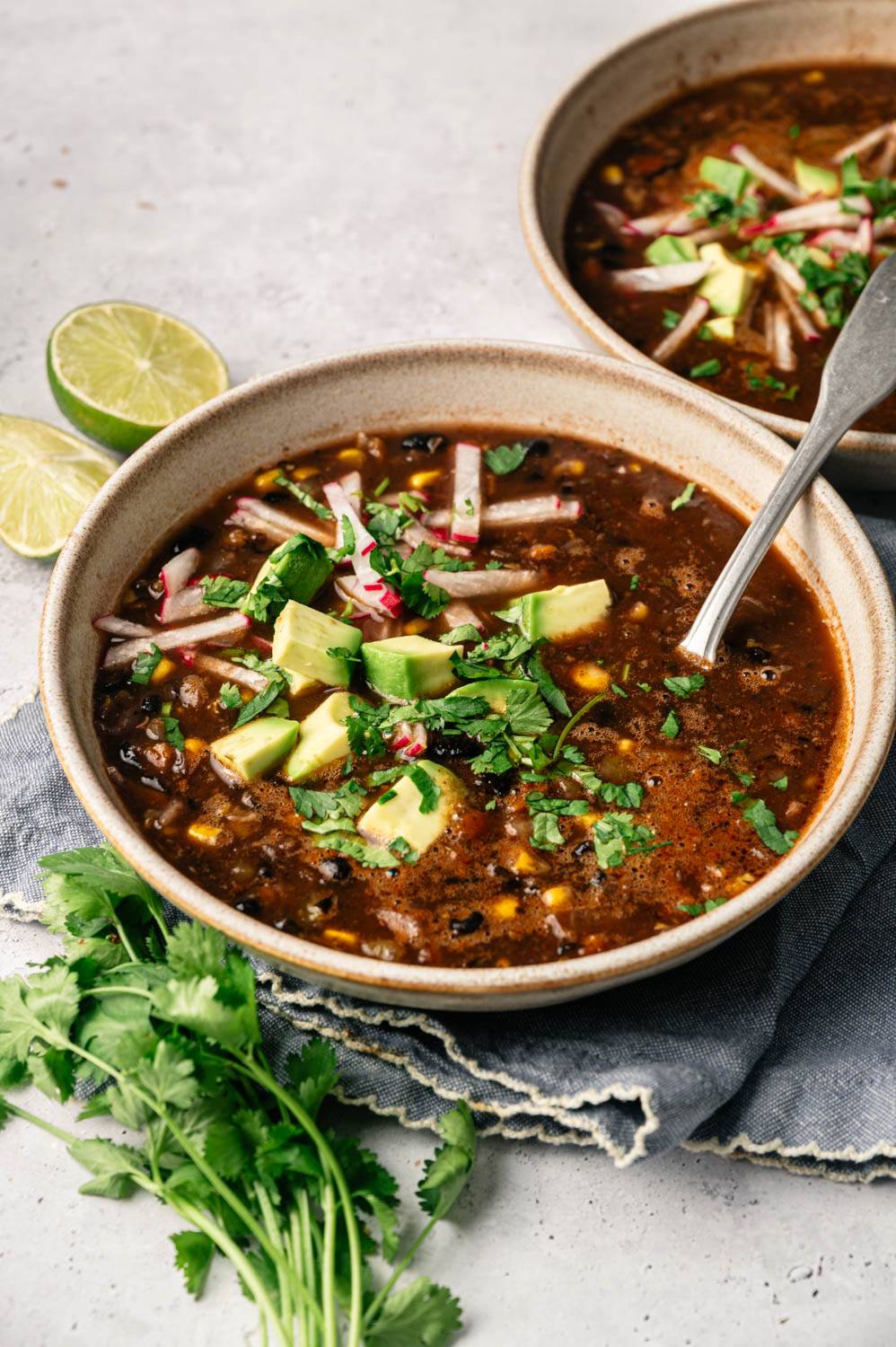 A bowl of hearty black bean soup garnished with avocado, radish, and cilantro, placed on a gray cloth. Lime halves and cilantro sprigs are nearby.