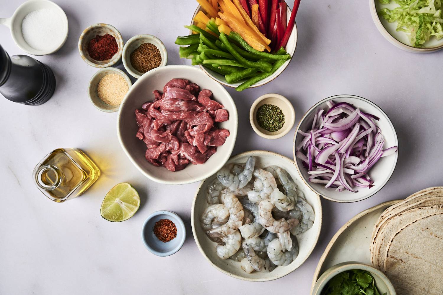 A variety of fresh ingredients for cooking, including shrimp, beef, colorful peppers, onions, and spices, arranged on a light surface.