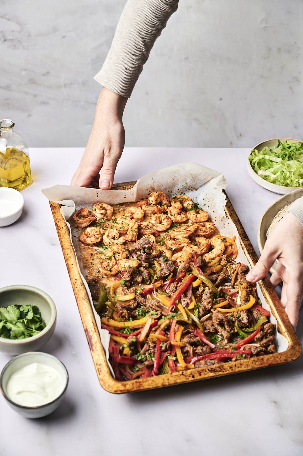 A person lifts a tray of shrimp and seasoned beef with colorful bell peppers, garnished with herbs, alongside bowls of salad and sauces.