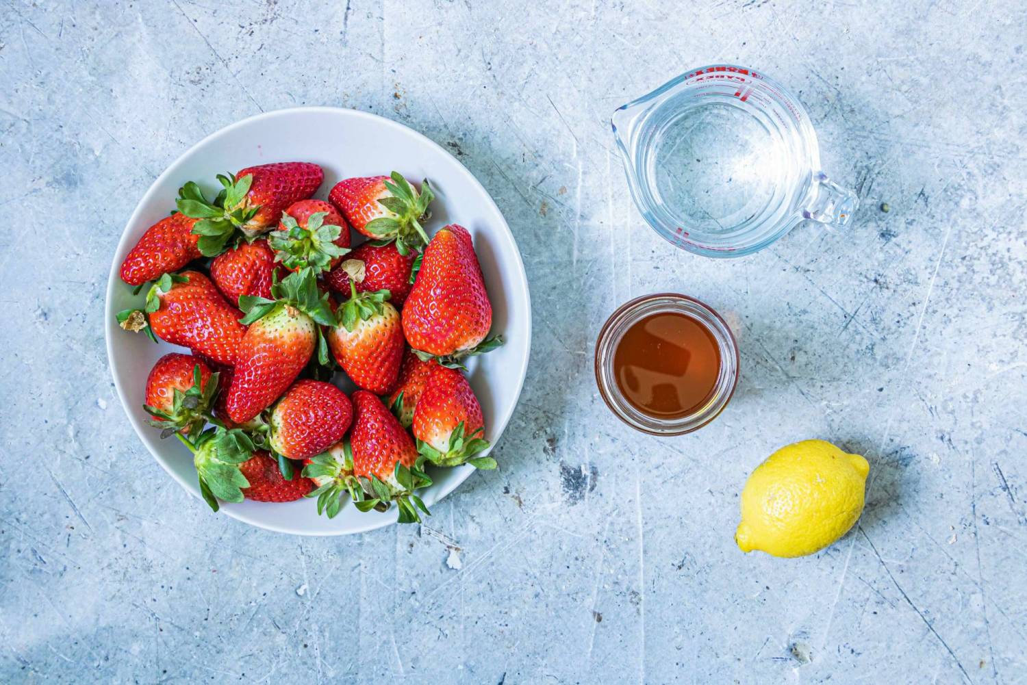 A plate of fresh strawberries, a measuring cup of water, a jar of honey, and a lemon on a light gray surface.