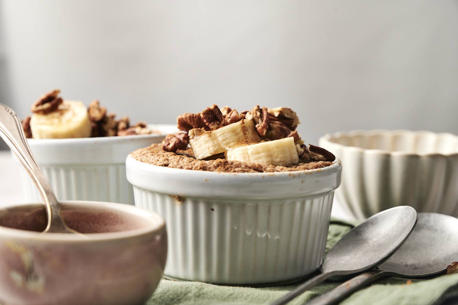 Close-up of a ramekin filled with baked protein oats, topped with banana slices, pecans, and cinnamon. Additional ramekin and bowl in soft focus in the background, with spoons and a green napkin in the foreground.
