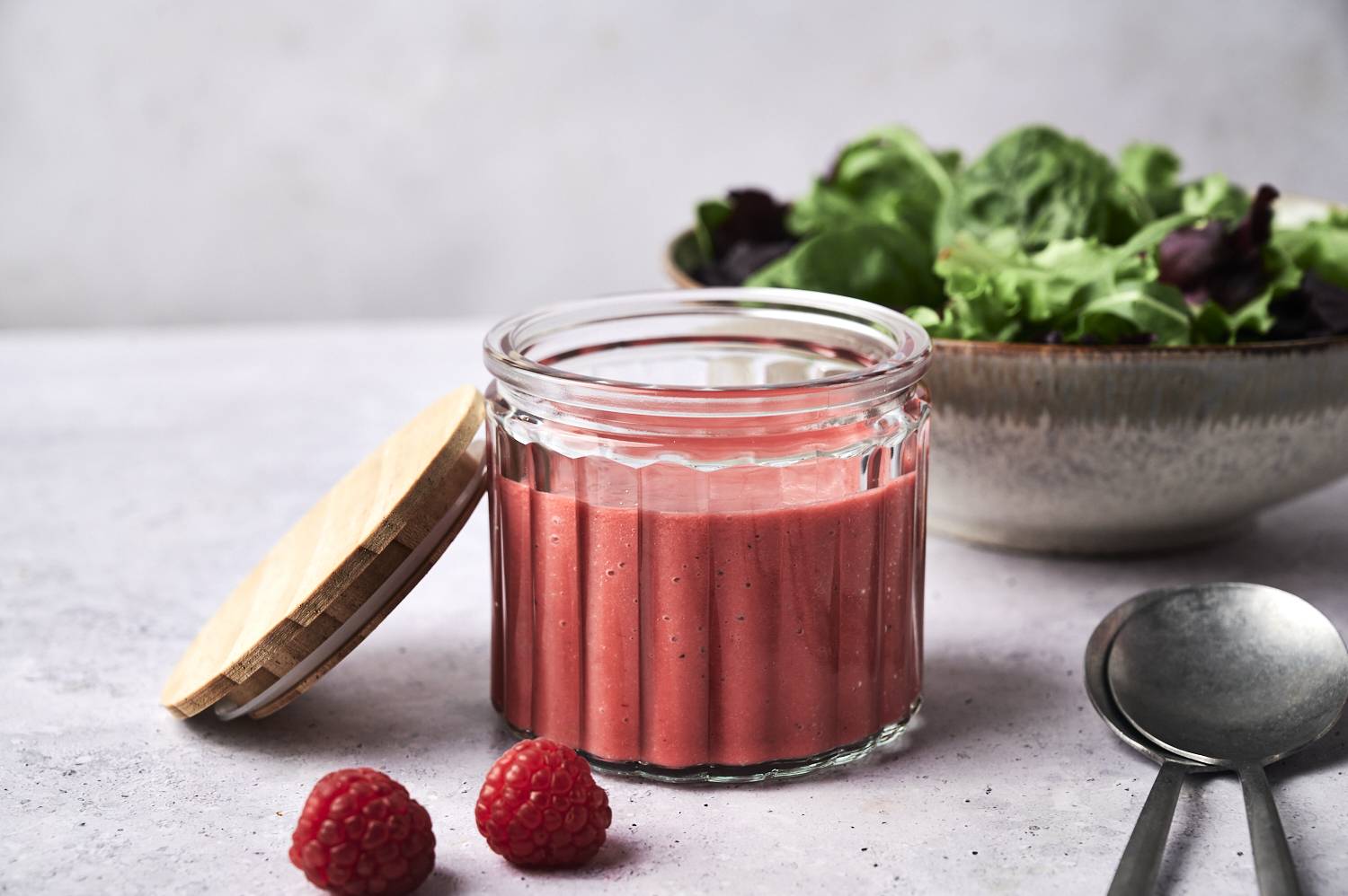 A jar of vibrant pink raspberry dressing sits beside fresh salad greens and two silver spoons on a textured grey surface.