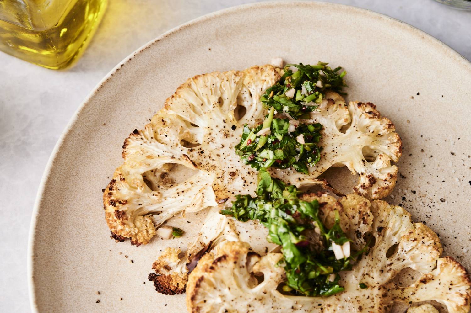 Close-up of golden roasted cauliflower steaks topped with fresh chimichurri sauce on a beige plate.
