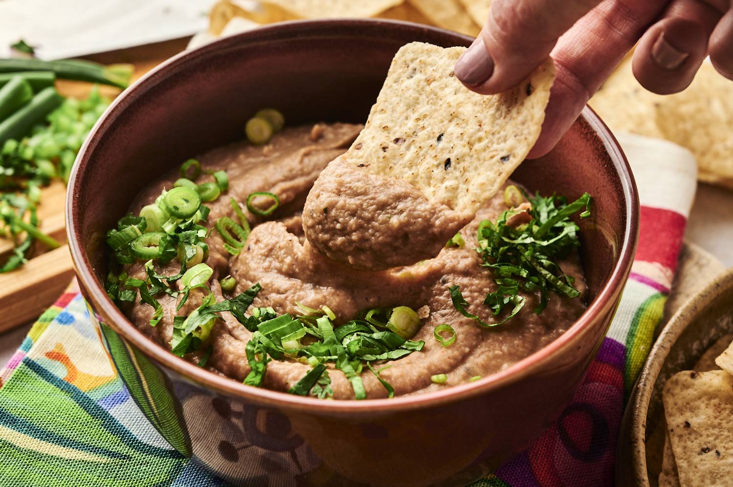 A tortilla chip is dipped into a bowl of creamy refried beans topped with fresh green onions and cilantro, ready to enjoy.