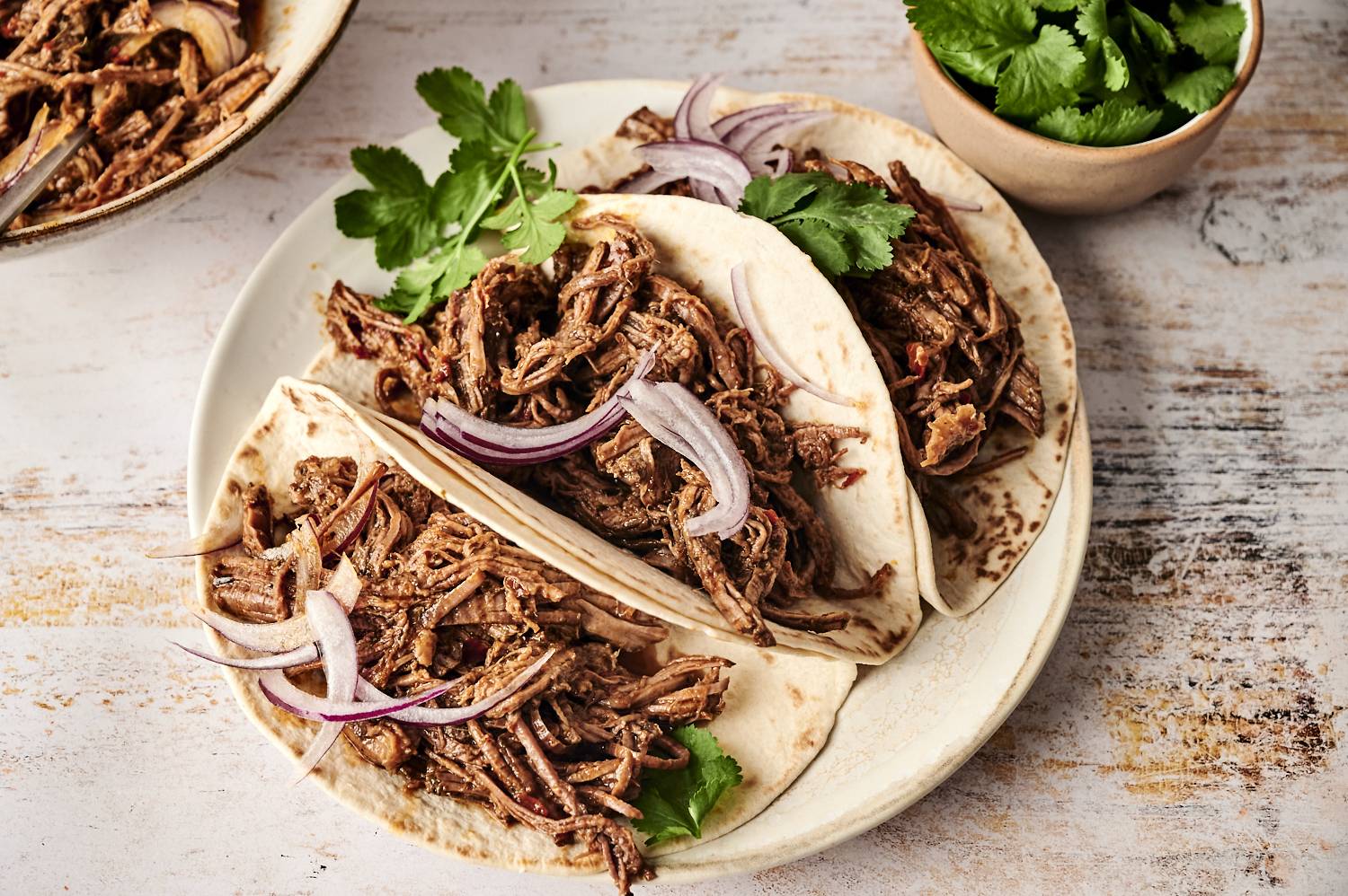 Soft flour tortillas are filled with tender, shredded Mexican beef adobo, topped with sliced red onions and fresh cilantro, ready to serve.