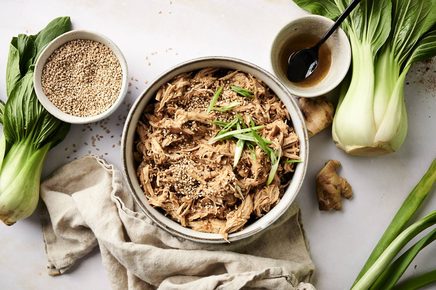 Overhead view of shredded slow-cooked sesame ginger chicken in a bowl garnished with sesame seeds and green onions, surrounded by fresh bok choy, ginger, sesame seeds, and a bowl of sauce on a light countertop