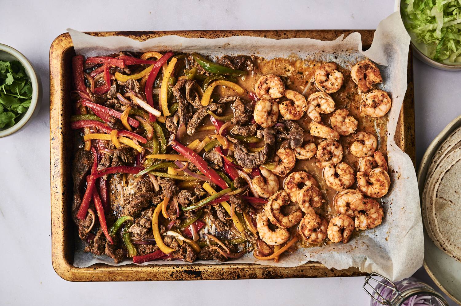 A baking tray with seasoned shrimp and beef mixed with colorful bell peppers, surrounded by fresh cilantro and chopped lettuce.