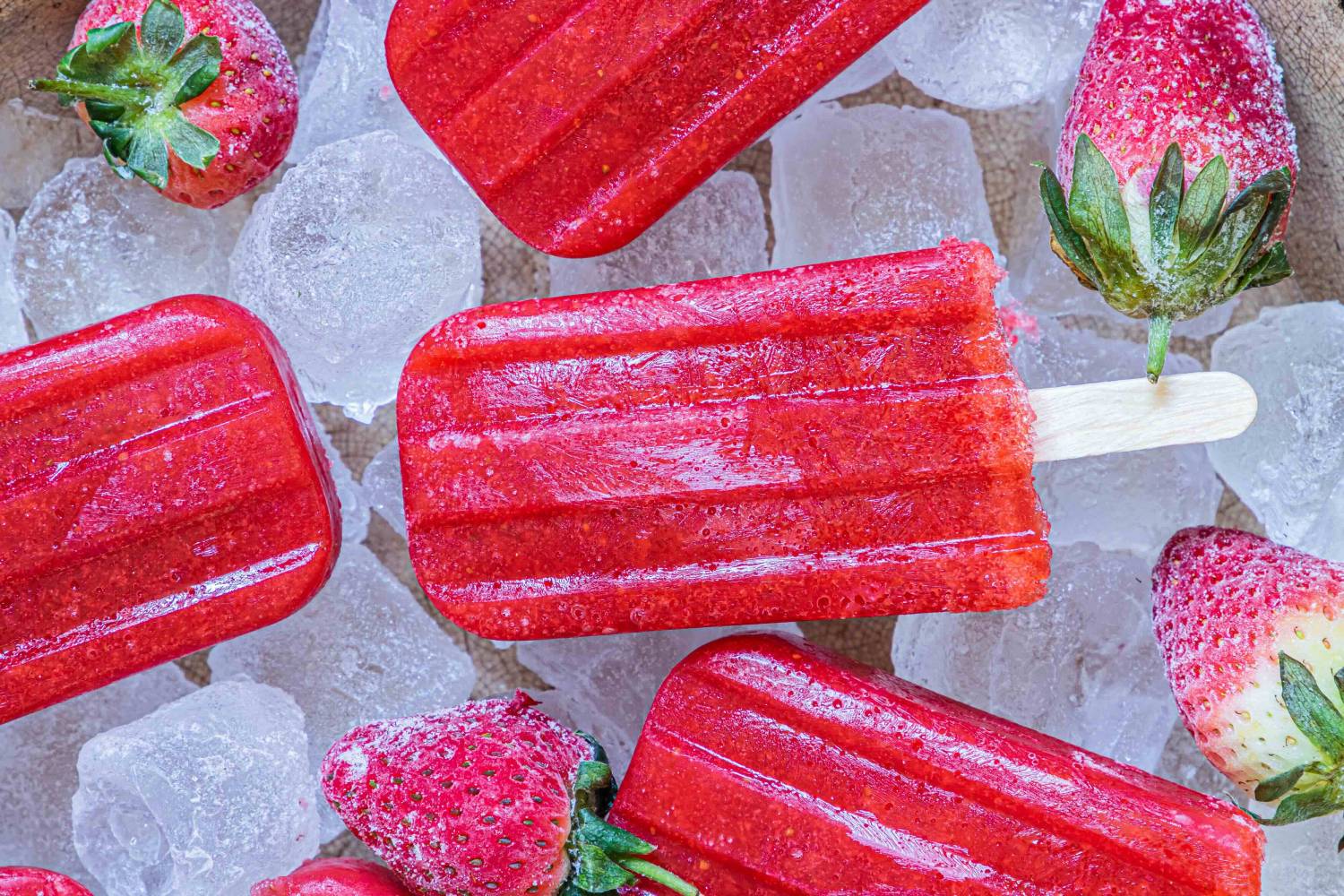Bright red strawberry popsicles on ice, surrounded by fresh strawberries and a yellow flower, evoking a refreshing summer treat.