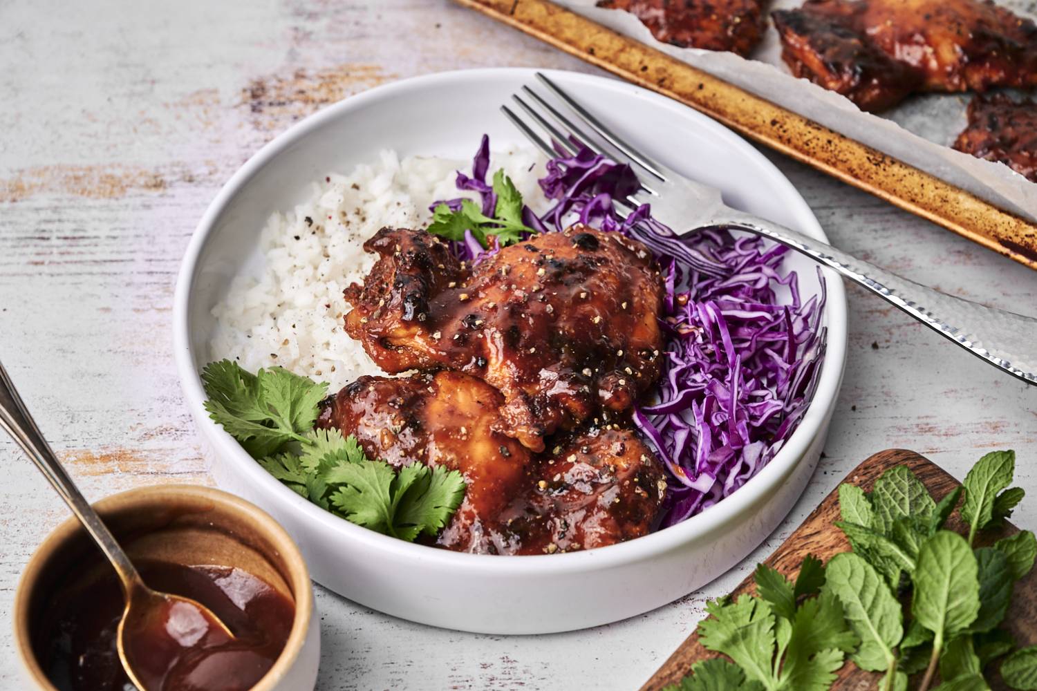 A white bowl filled with baked BBQ chicken thighs glazed in a rich sauce, served alongside fluffy white rice, shredded purple cabbage, and fresh cilantro, with a fork resting on the side.
