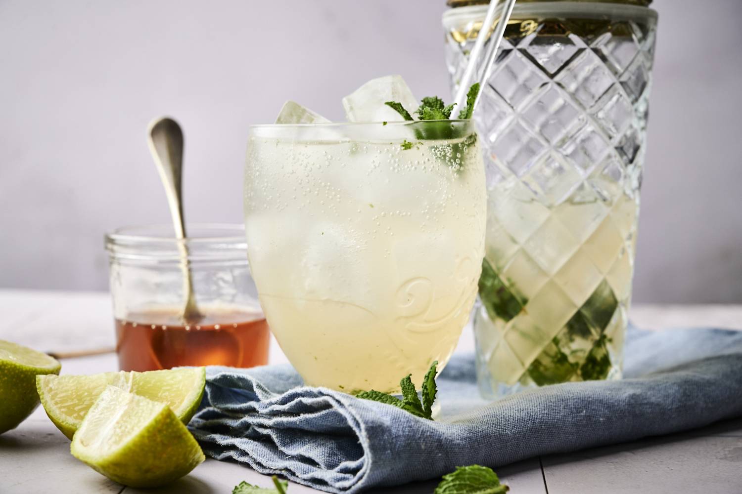 A refreshing drink with mint and ice, next to a jar of honey and lime wedges on a blue napkin.