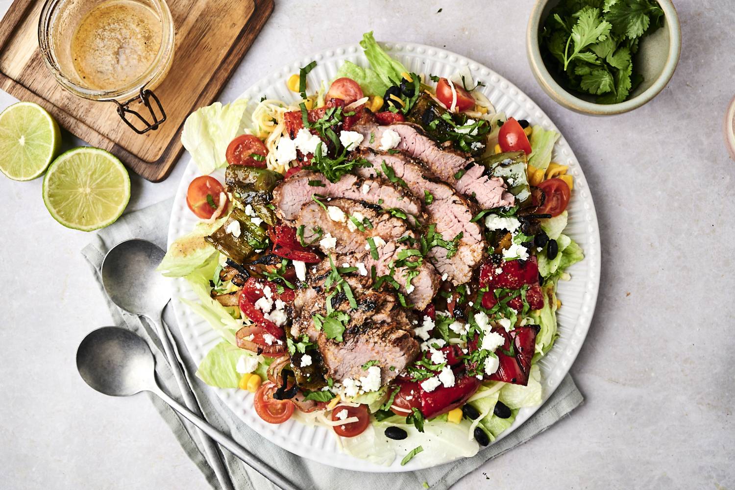 A vibrant salad topped with sliced grilled beef, assorted veggies, feta cheese, and cilantro, accompanied by lime halves and dressing.