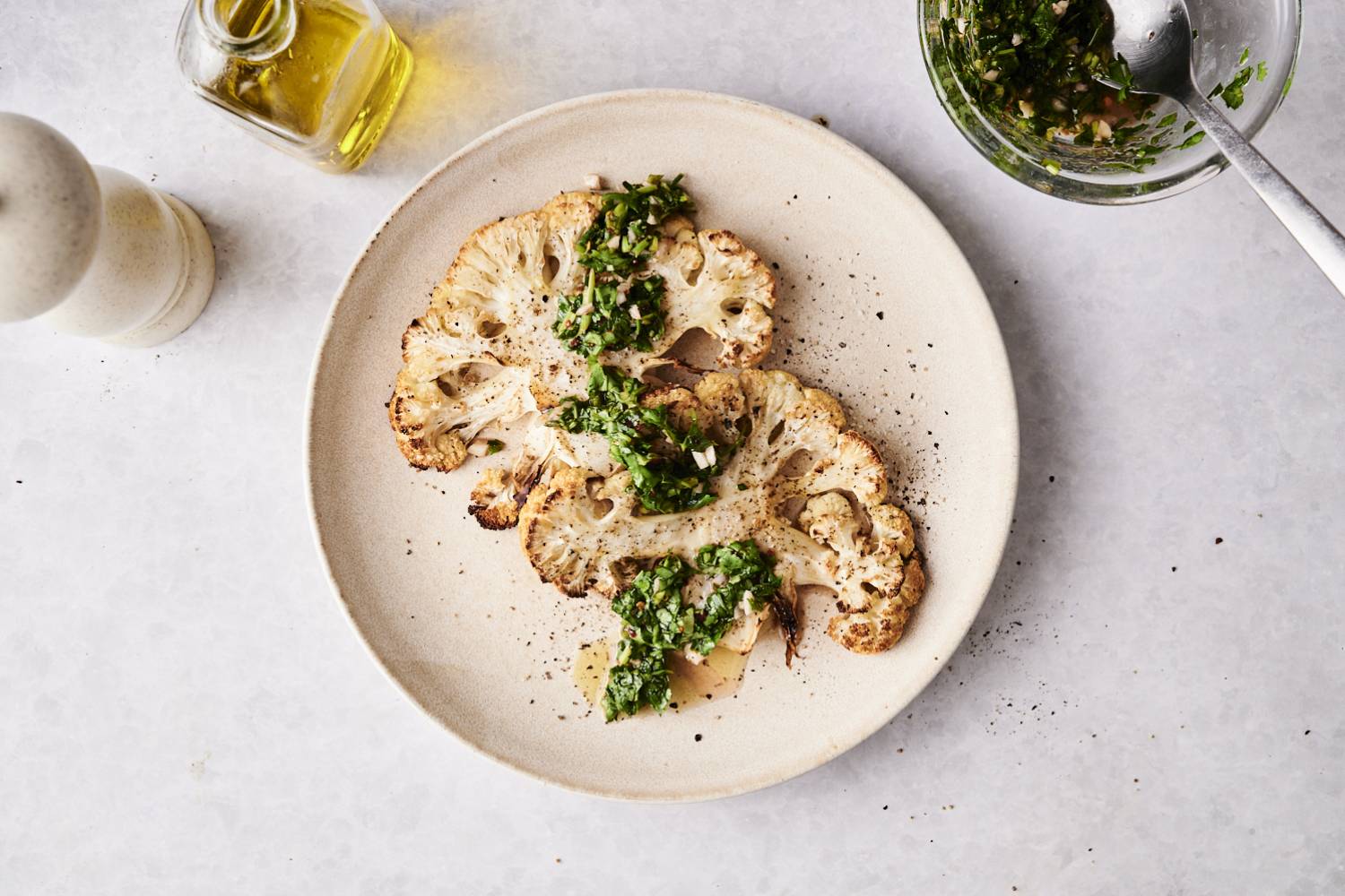Overhead view of roasted cauliflower steaks garnished with chimichurri sauce, served on a plate with olive oil and pepper mill nearby.