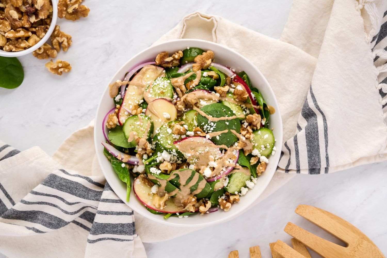 Spinach salad with creamy balsamic vinaigrette, goat cheese, apples, and walnuts in a white bowl with wooden salad tongs.