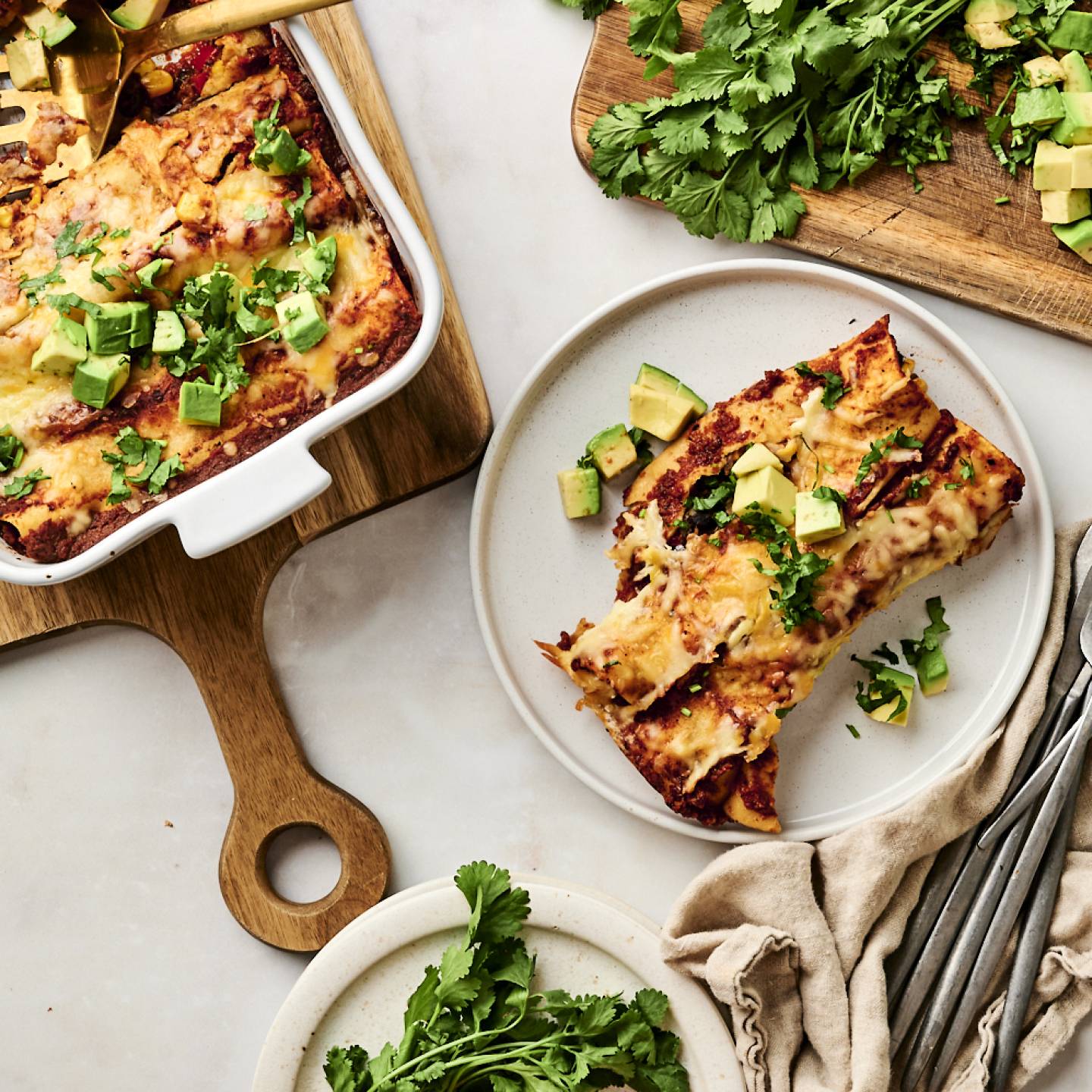 Cheesy black bean enchiladas topped with diced avocado and fresh cilantro, served on a white plate with a side of extra herbs on a wooden cutting board.