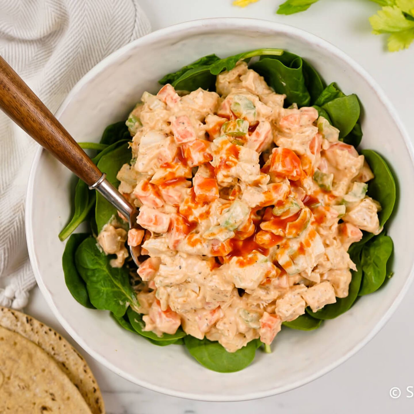 Buffalo chicken salad with fresh spinach, a whole wheat wrap, and a spoon in a bowl.