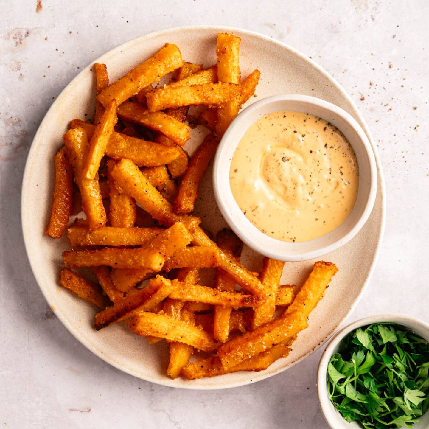 Plate of crispy golden fries with a creamy dipping sauce on the side. A small bowl of fresh green parsley is nearby on a light, textured surface.