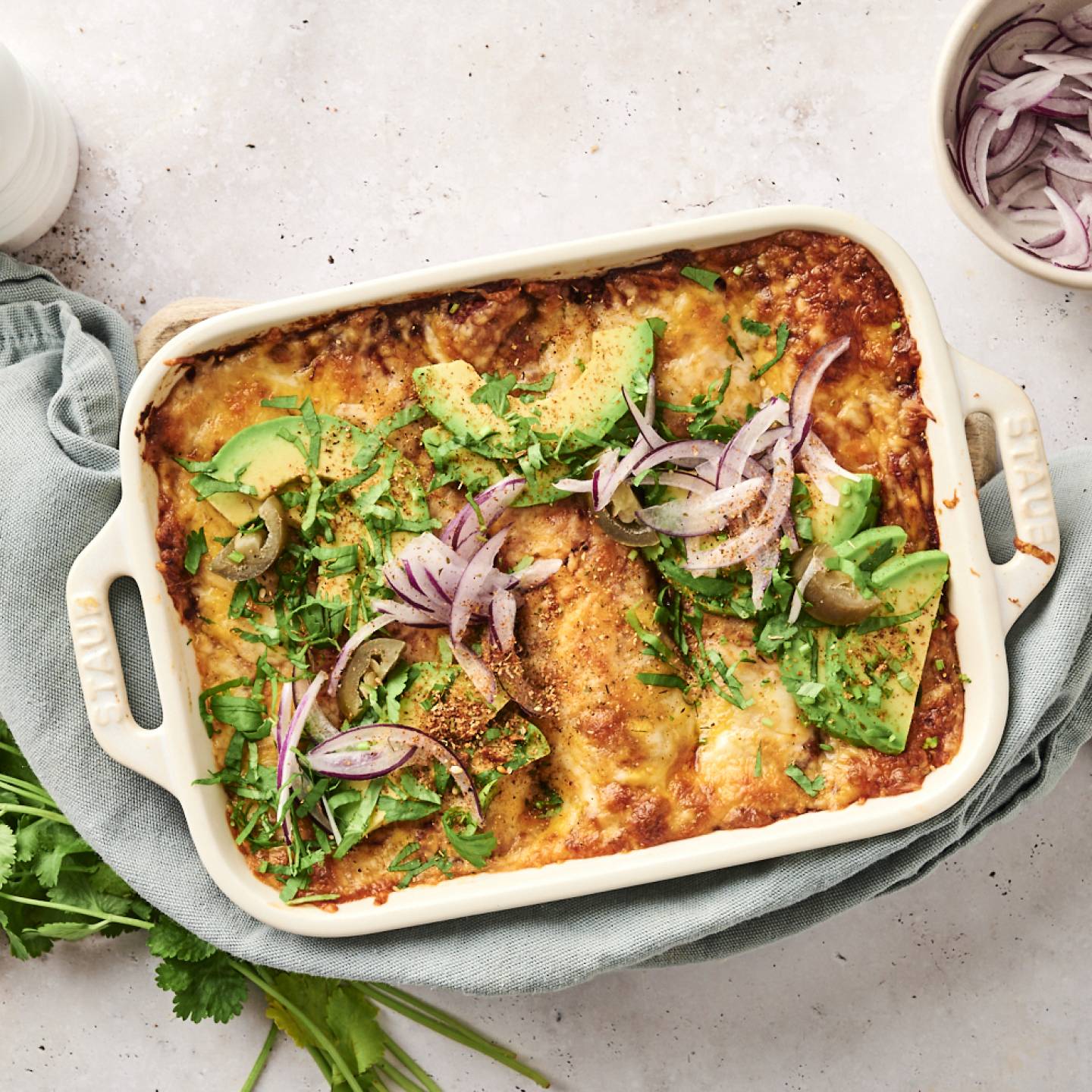 A close-up of cheesy chicken enchiladas being served from a casserole dish, topped with avocado, red onion, jalapeños, and fresh cilantro.