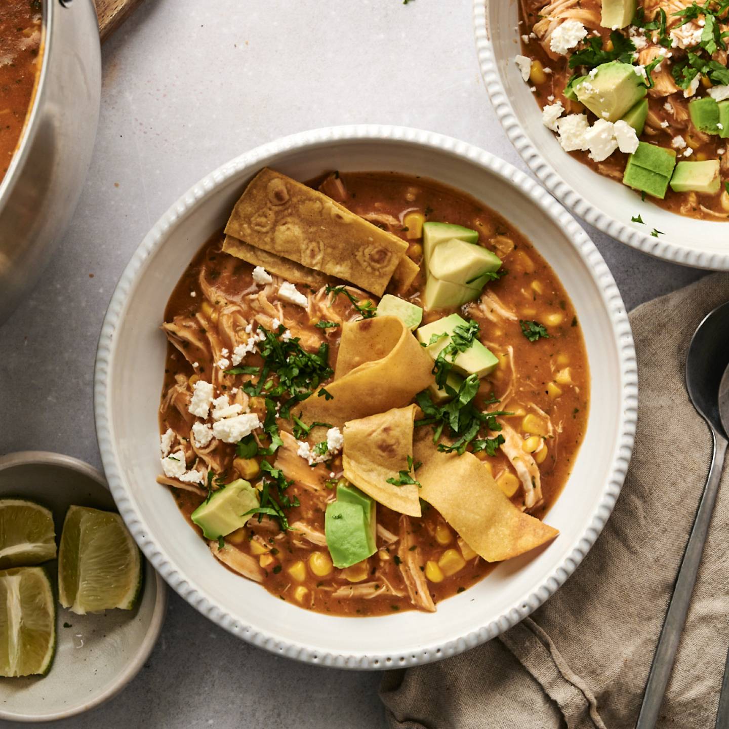 Top-down view of chicken tortilla soup served in white bowls, garnished with crispy tortilla strips, avocado, cilantro, and crumbled cheese, with lime wedges and a pot of soup in the background.