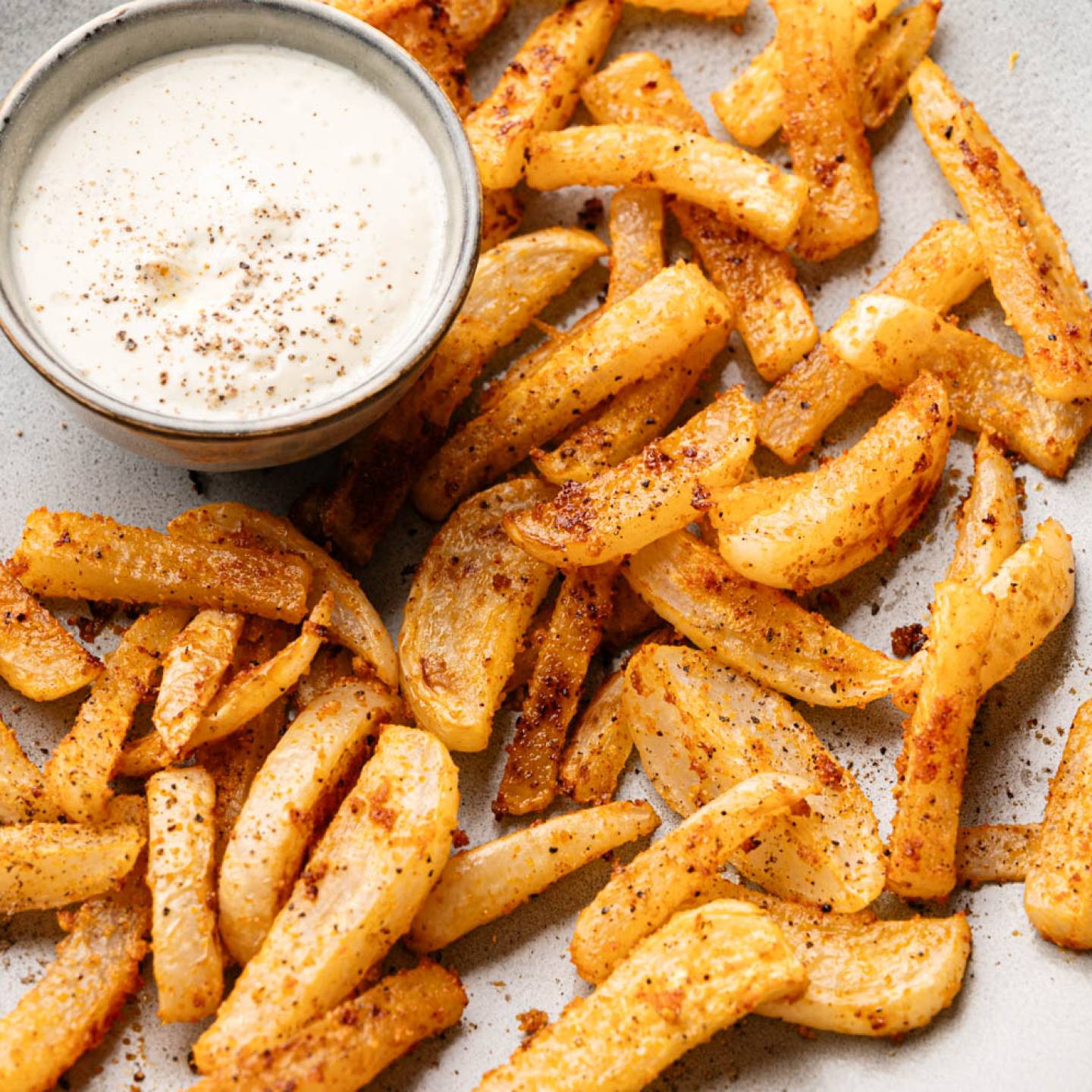 Crispy seasoned fries are arranged on a plate with a small bowl of creamy dipping sauce. The fries are golden and dusted with spices.