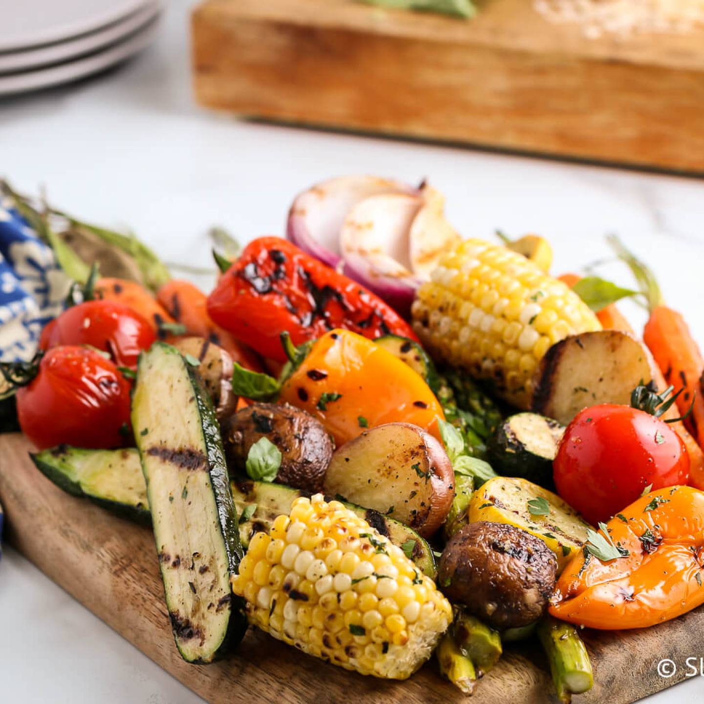 Grilled vegetables including zucchini, peppers, tomatoes, potatoes, and carrots on a wooden cutting board.