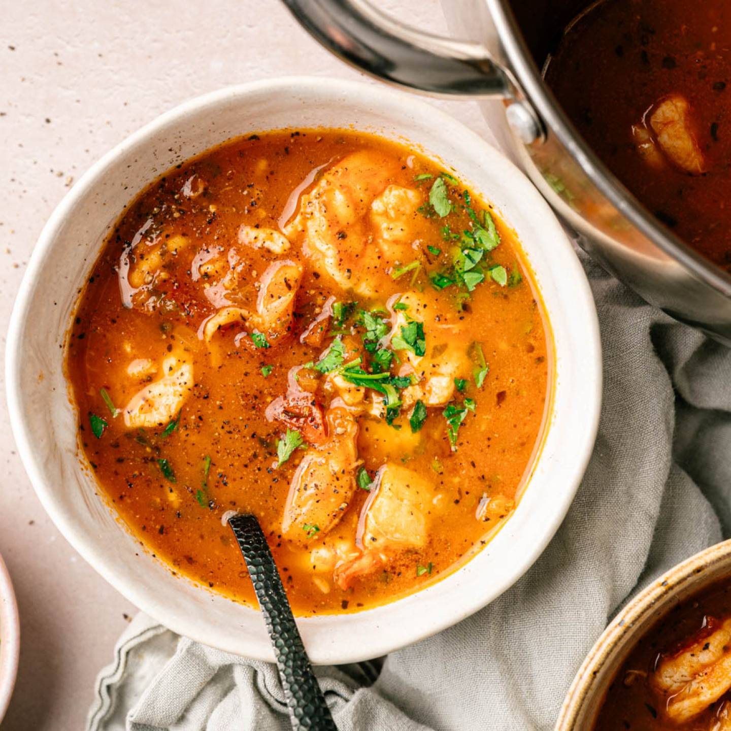 A bowl of hearty tomato soup with shrimp and herbs. The soup is a rich, warm reddish color, garnished with chopped parsley, next to a pot. Cozy and appetizing.