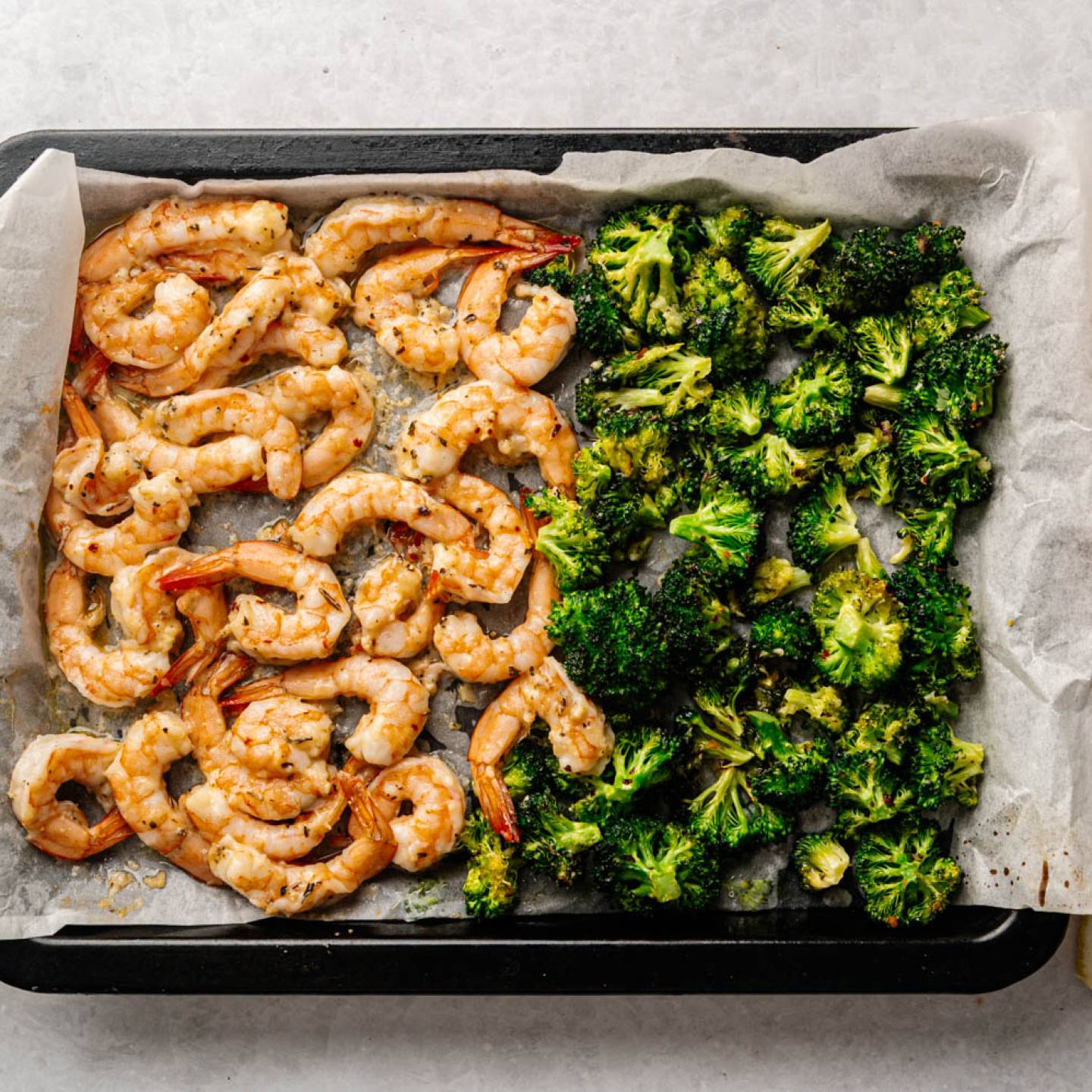 Roasted shrimp and broccoli on a baking sheet lined with parchment paper. Olive oil, lemon wedges, and a pepper mill are nearby. Bright and appetizing.