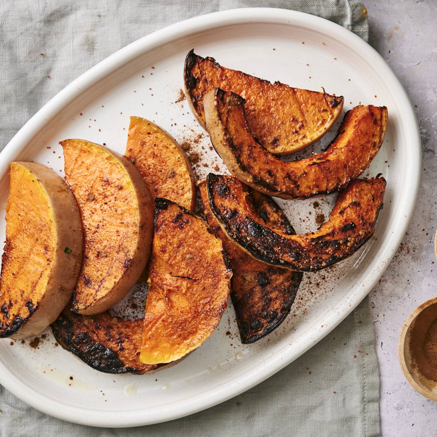 Grilled butternut squash slices on a white platter, garnished with spices, alongside two small bowls of paprika and cinnamon.