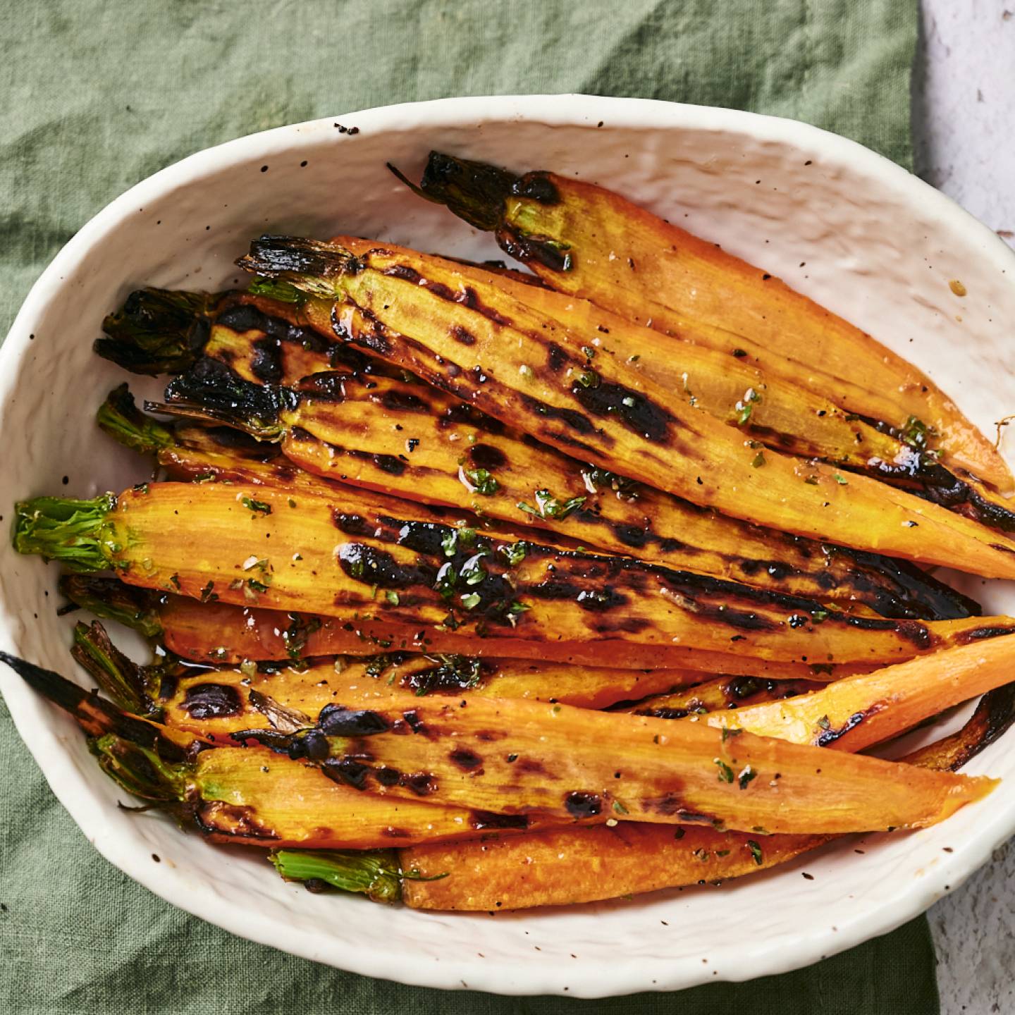 A white bowl filled with charred orange carrots topped with herbs, resting on a green cloth and surrounded by a textured surface.
