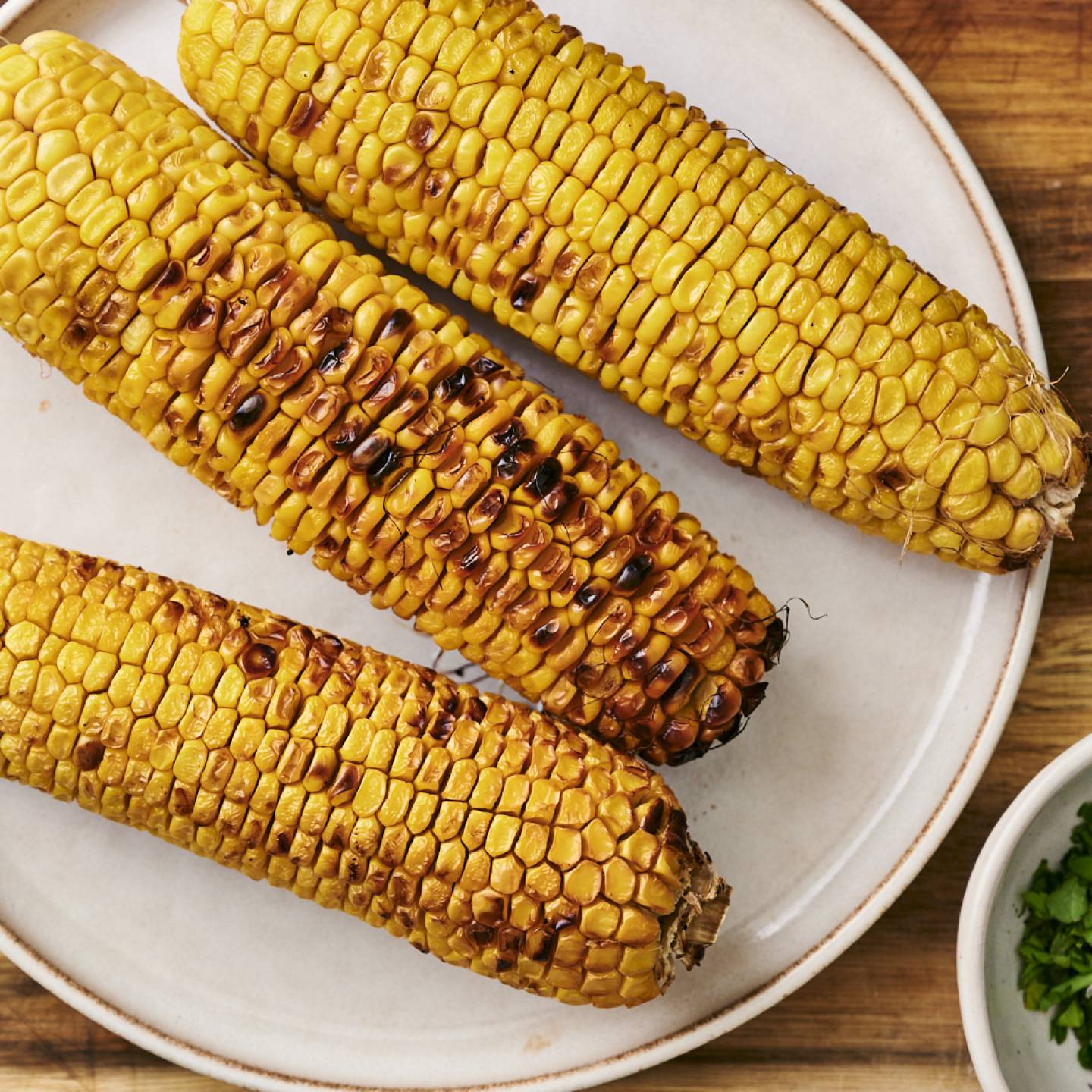 Three grilled corn cobs with golden kernels sit on a white plate beside a small bowl of chopped green herbs.