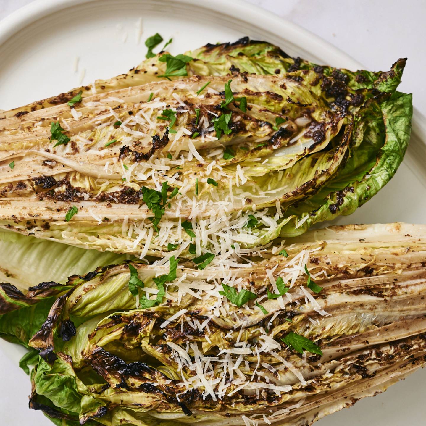 Grilled romaine lettuce halves topped with grated Parmesan, herbs, and black pepper, served with a lemon slice on a white plate.