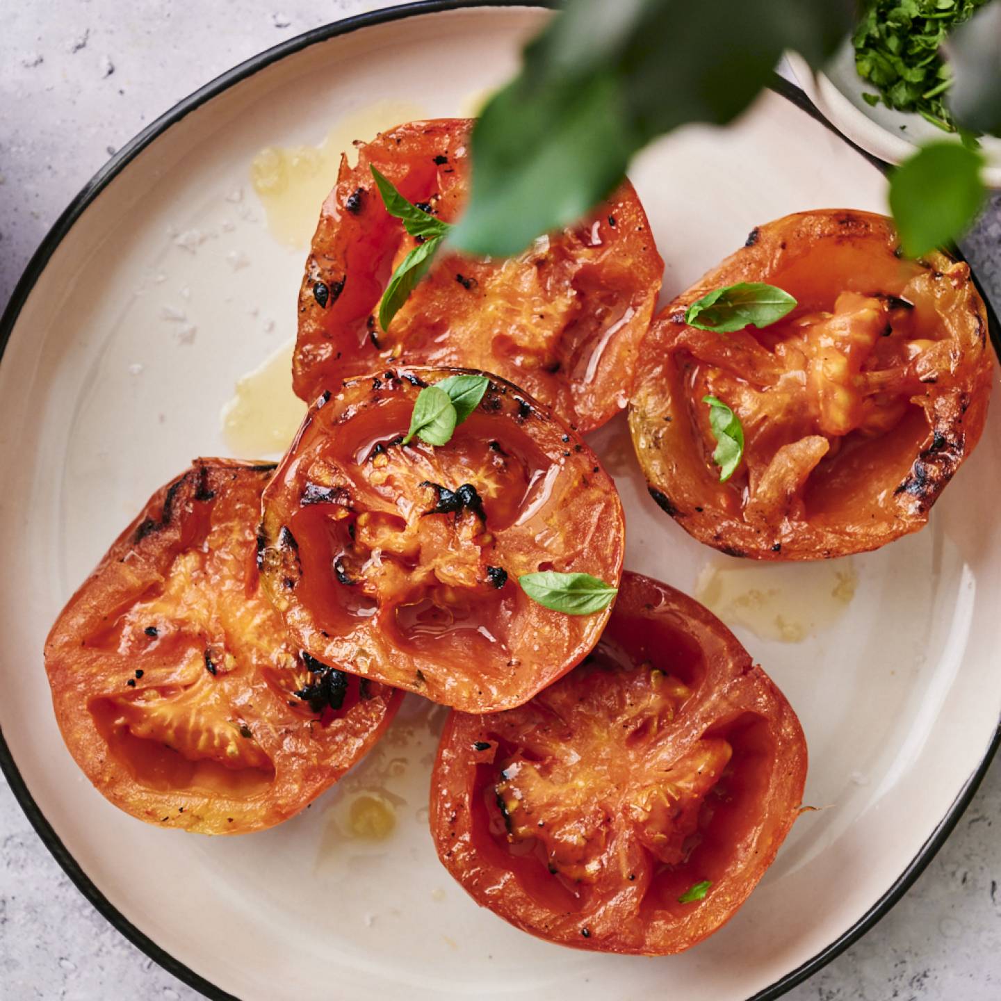 Grilled tomatoes topped with fresh basil, drizzled with olive oil, served on a round plate alongside a bowl of broth.