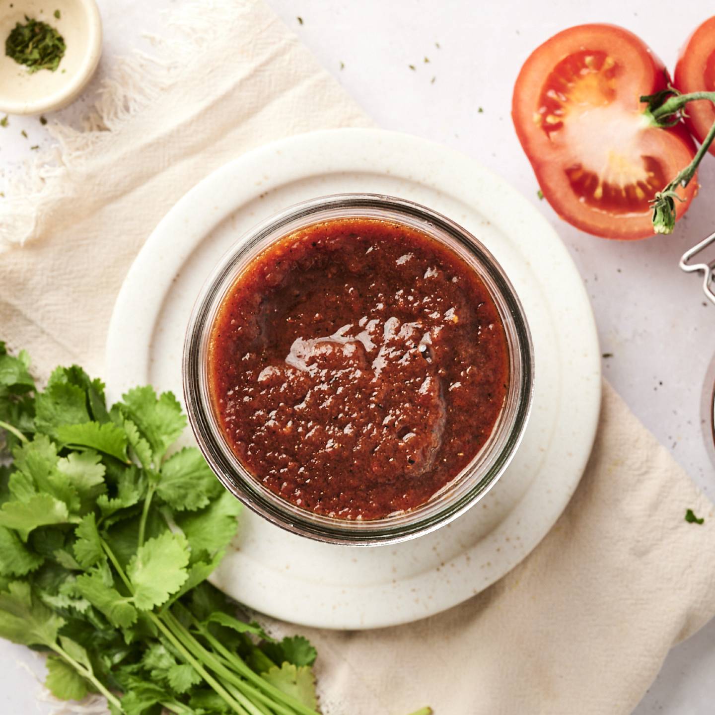 A jar of rich red enchilada sauce surrounded by fresh cilantro, tomatoes, and seasonings on a white backdrop.