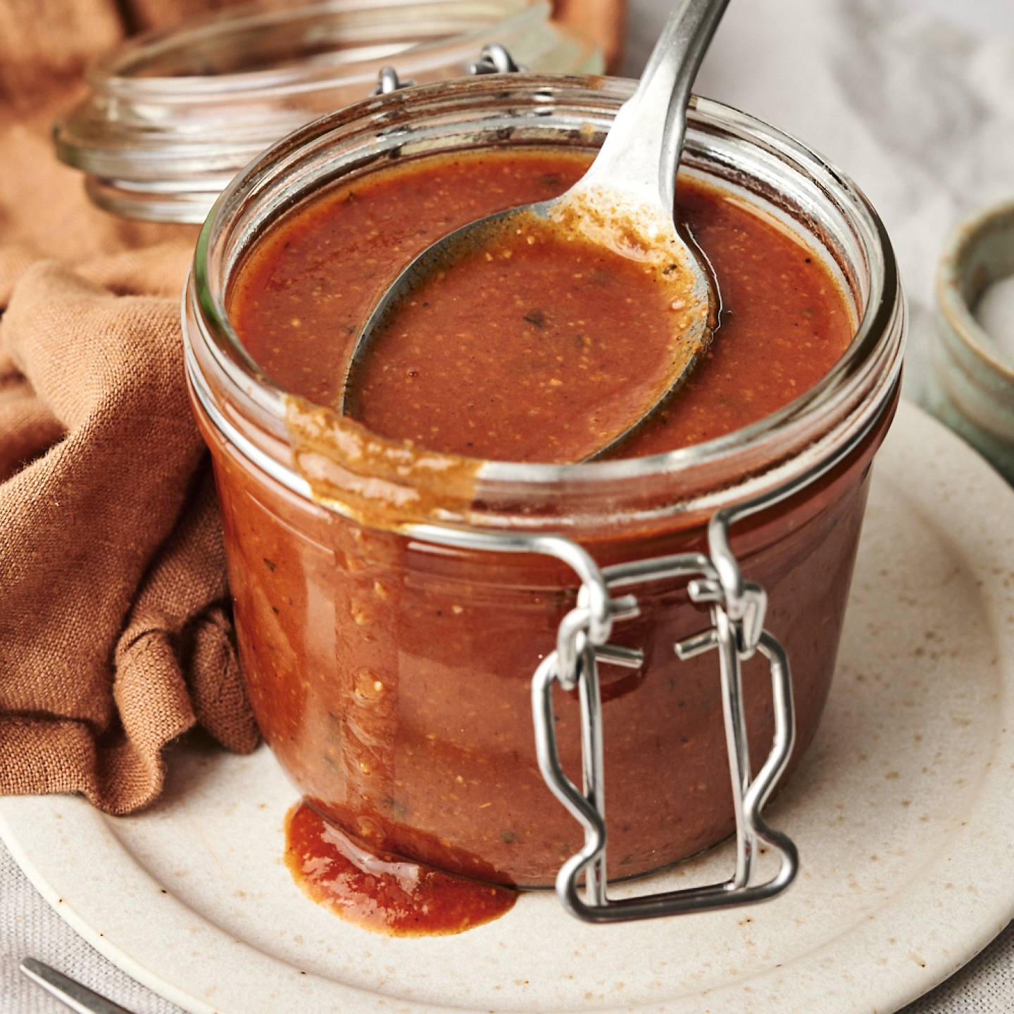 A glass jar filled with homemade enchilada sauce, with a spoon resting inside, placed on a ceramic plate with a rustic cloth and a small bowl of salt in the background.