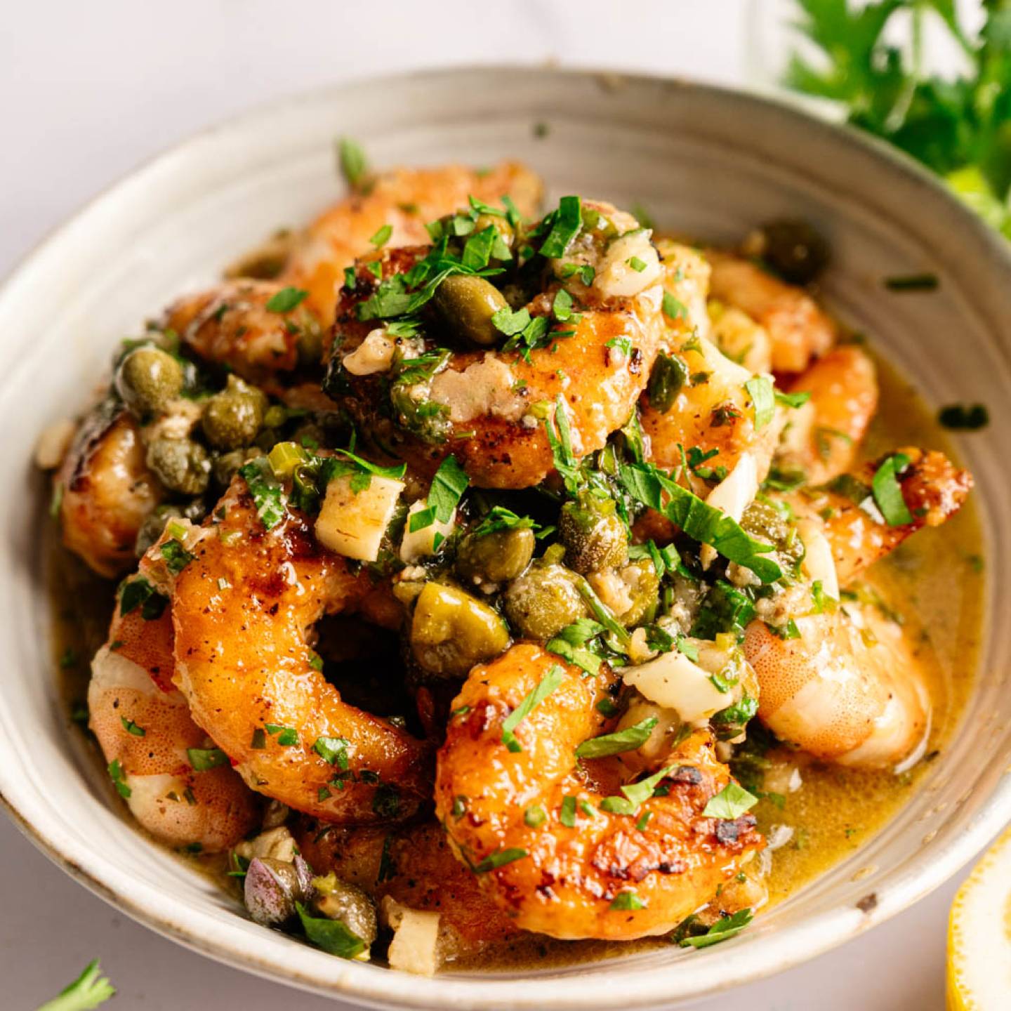 A bowl of golden-brown shrimp coated in a lemony garlic butter sauce, garnished with capers and fresh herbs. The setting conveys a fresh, appetizing tone.