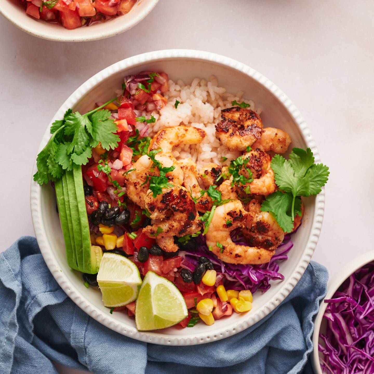 Shrimp taco bowls with cooked shrimp, avocado, rice, lime, cilantro, cabbage, corn, and pico de gallo in a white bowl.