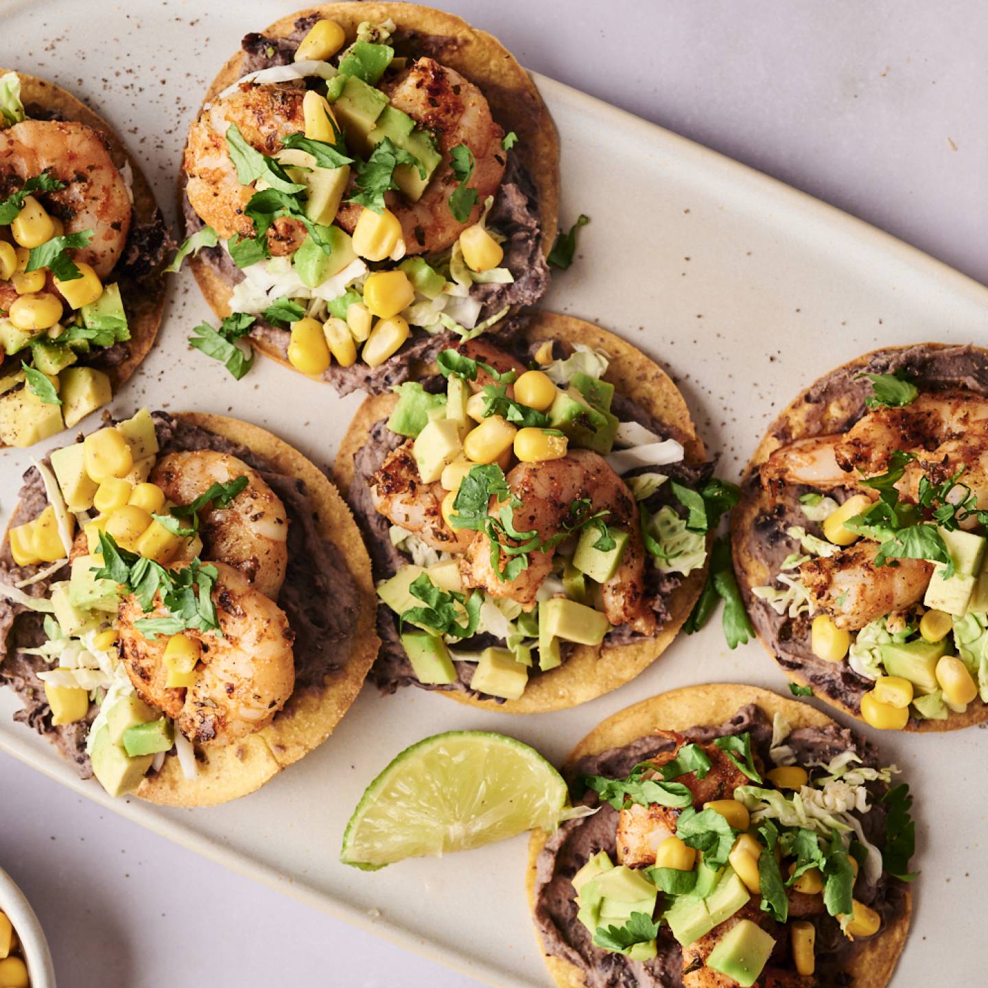 A plate of six shrimp tostadas with black beans, shredded cabbage, corn, avocado, and cilantro.