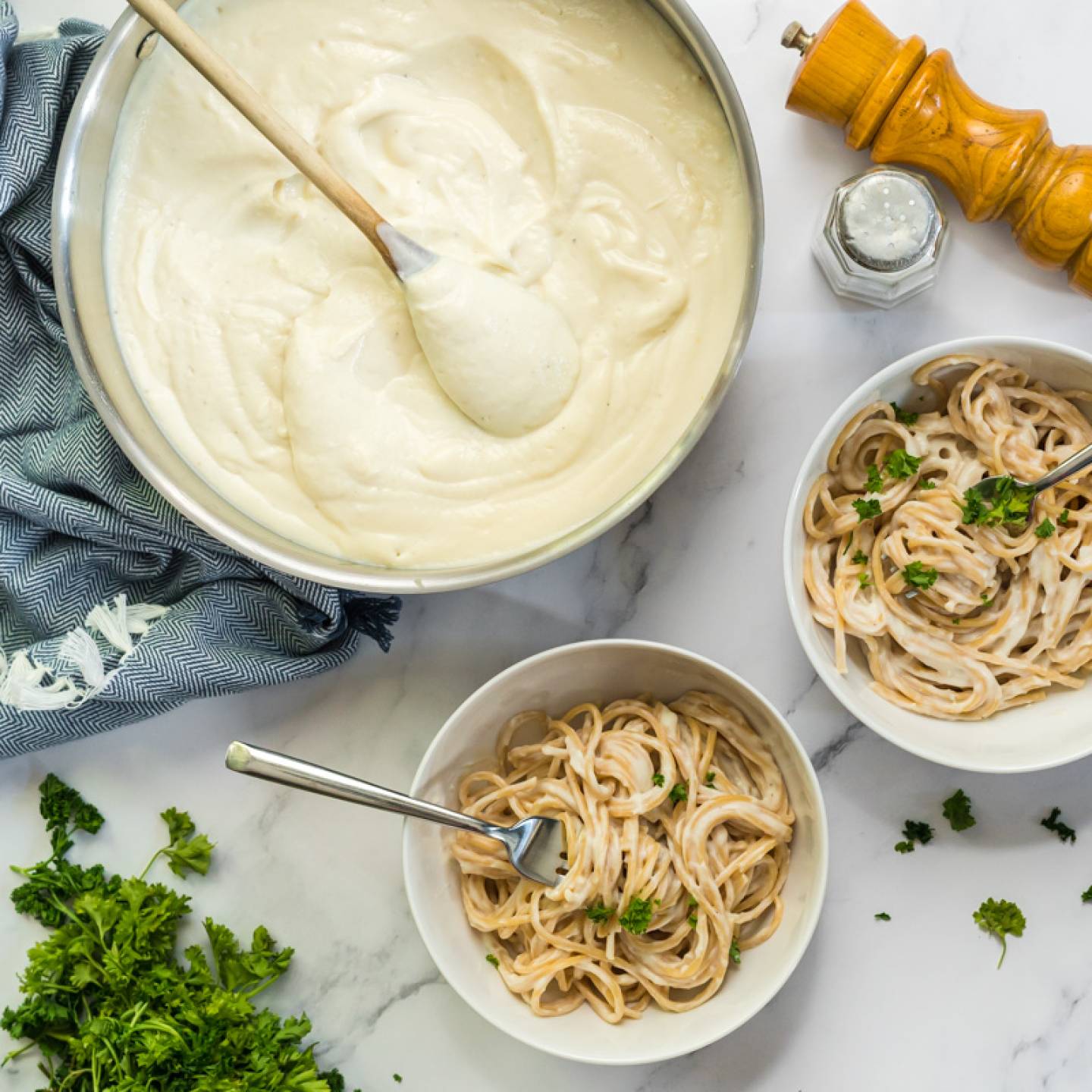 Skinny Alfredo sauce in a large pan with a wooden spoon and served in two bowls with spaghetti and parsley.