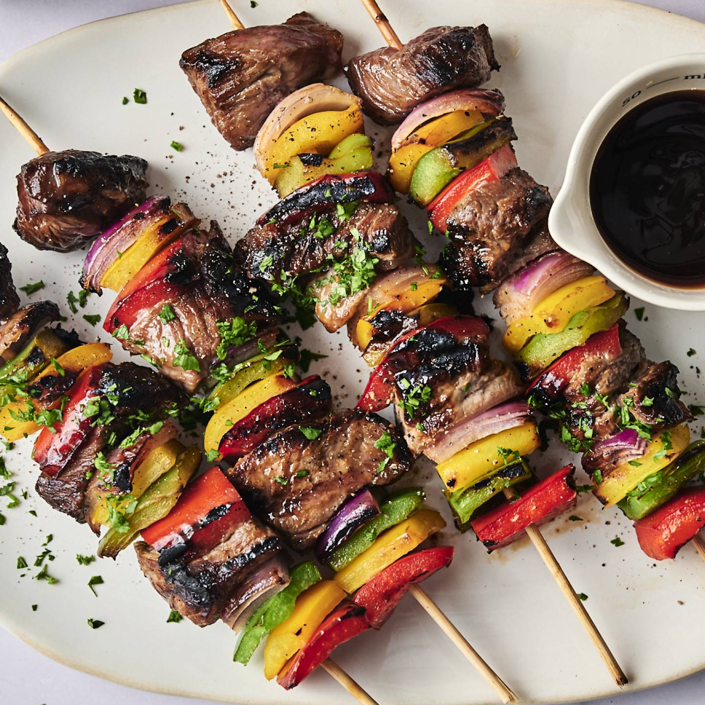 Grilled kebabs featuring marinated beef and colorful vegetables, served with a small bowl of dipping sauce on a white plate.